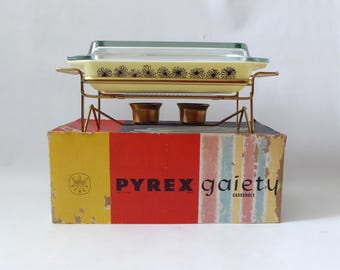 May include: A rectangular yellow Pyrex Gaiety casserole dish with a clear glass lid, featuring a black daisy pattern. The dish rests on a gold-toned metal warming stand with two candle holders, all positioned atop its original cardboard box.