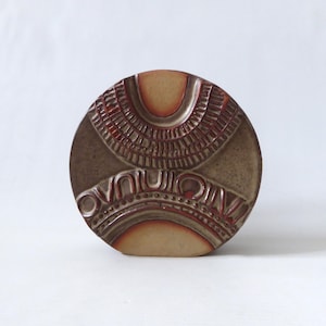 May include: A circular ceramic vase featuring a matte tan finish with deep reddish-brown accents. The surface is decorated with embossed, repeating geometric patterns, including vertical lines and curved arches that create a textured relief.
