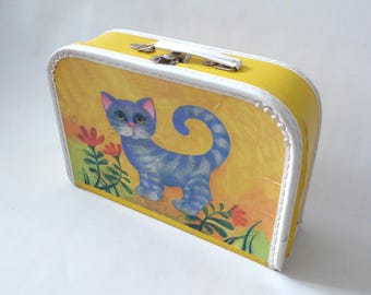 May include: A small yellow rectangular suitcase with a white trim and a top handle. The front panel features a blue striped cat illustration standing among red flowers against a yellow background. The case has silver metal hardware.