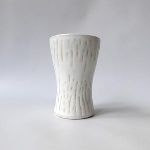 May include: A white ceramic vase with a textured, vertical pattern. The vase has a slightly hourglass shape, wider at the top and bottom, and narrower in the middle. The surface has a matte finish.