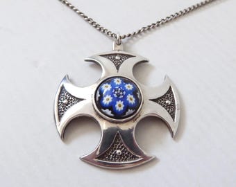 May include: A silver-toned cross pendant on a thin chain. The cross features a central circular inlay of blue millefiori glass with white and yellow flower patterns. The metal arms of the cross have a smooth finish with textured, dark recessed areas.