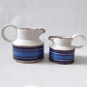May include: Two ceramic jugs with a white speckled upper section and a lower section featuring horizontal bands of dark blue, medium blue, and light blue. Each jug has a rounded handle and a spout, with brown edges along the rim.