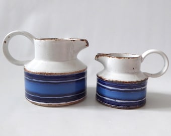May include: Two ceramic jugs with a white speckled upper section and a lower section featuring horizontal bands of dark blue, medium blue, and light blue. Each jug has a rounded handle and a spout, with brown edges along the rim.