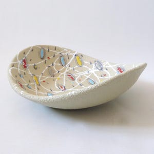 May include: A beige ceramic bowl with a textured exterior and an asymmetrical, leaf-like shape. The interior features a white, intersecting line pattern overlaid with small, colourful oval shapes in red, yellow, and light blue throughout.