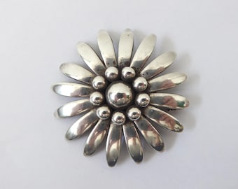 Könnte beinhalten: Eine kreisförmige silberne Brosche in Blumenform mit einer großen Zentralkugel, die von einem Ring aus kleineren Kugeln und länglichen, glatten, blütenblattartigen Silbersegmenten umgeben ist, die strahlenförmig nach außen verlaufen.