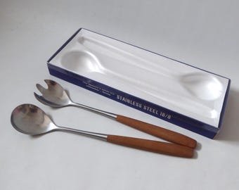 Könnte beinhalten: Ein Salatbesteck bestehend aus einer Gabel und einem Löffel mit Edelstahlköpfen und braunen Holzgriffen. Das Set wird in einer rechteckigen blau-weißen Schachtel mit der Aufschrift STAINLESS STEEL 18/8 und geformten weißen Kunststoffeinsätzen präsentiert.