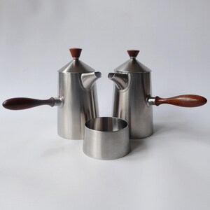 May include: A set of three stainless steel coffee or tea pots with wooden handles. The pots have a rounded, conical shape with a small spout and a lid with a wooden knob. The set includes a small, round, stainless steel creamer.