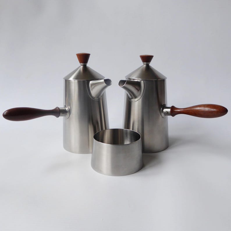 May include: A set of three stainless steel coffee or tea pots with wooden handles. The pots have a rounded, conical shape with a small spout and a lid with a wooden knob. The set includes a small, round, stainless steel creamer.