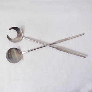 May include: Two crossed stainless steel serving utensils against a white backdrop. One has a circular bowl, the other a crescent-shaped head. Both have long, slender handles, likely for serving or cooking purposes in a kitchen setting.