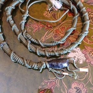 May include: A coiled, olive green rope with a black handle and white tassel. The rope is made of tightly woven material and appears to be handmade. The handle has a dark, glossy finish. The item is displayed on a floral patterned surface.