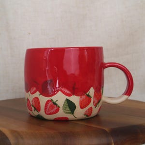 May include: A red ceramic mug with a white handle and a strawberry design. The mug has a glossy finish and a pattern of strawberries and green leaves around the base. The mug is on a wooden surface.