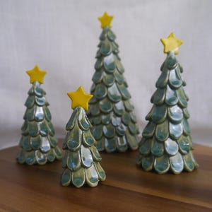 Könnte beinhalten: Vier keramische Weihnachtsbäume mit glänzender, blaugrüner Glasur und gelben Sternen. Die Bäume sind unterschiedlich groß und auf einer Holzoberfläche angeordnet, was eine festliche Weihnachtsdekoration ergibt. Die Bäume haben ein strukturiertes, schuppenartiges Muster.