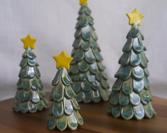 Op de afbeelding: Vier keramische kerstbomen met een glanzend, blauwgroen glazuur en gele sterren. De bomen variëren in grootte en zijn geplaatst op een houten oppervlak, wat een feestelijke kerstdisplay creëert. De bomen hebben een textuur met schubben.