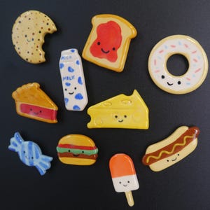 May include: A selection of food-themed ceramic magnets. The collection includes a cookie, toast with jam, a donut, a slice of pie, a milk carton, a cheese wedge, a piece of candy, a burger, a popsicle, and a hot dog. Each item has a smiling face.