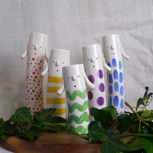 May include: Five white ceramic vases with different coloured patterns. The vases are shaped like people with arms and faces. The patterns include red polka dots, yellow stripes, green waves, purple polka dots, and blue polka dots.