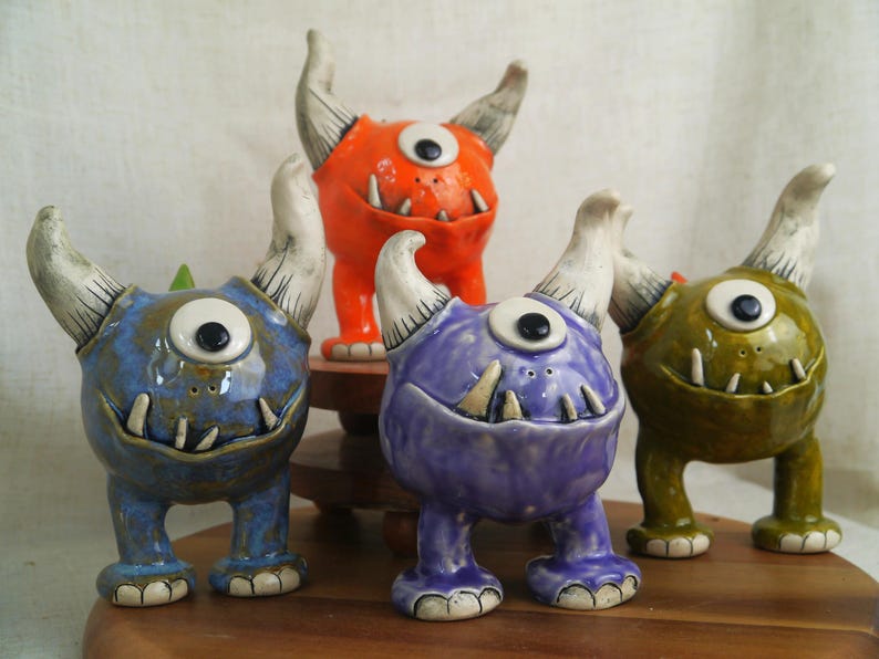 May include: A collection of ceramic monster figurines in various colors: blue, orange, purple, and green. Each has one eye, horns, and a toothy grin. The figurines are displayed on a wooden surface.