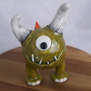 May include: A whimsical ceramic monster figurine in shades of olive green, white, and orange. The creature has one large eye, two curved horns, and a row of small teeth. The figurine is standing on a wooden surface.