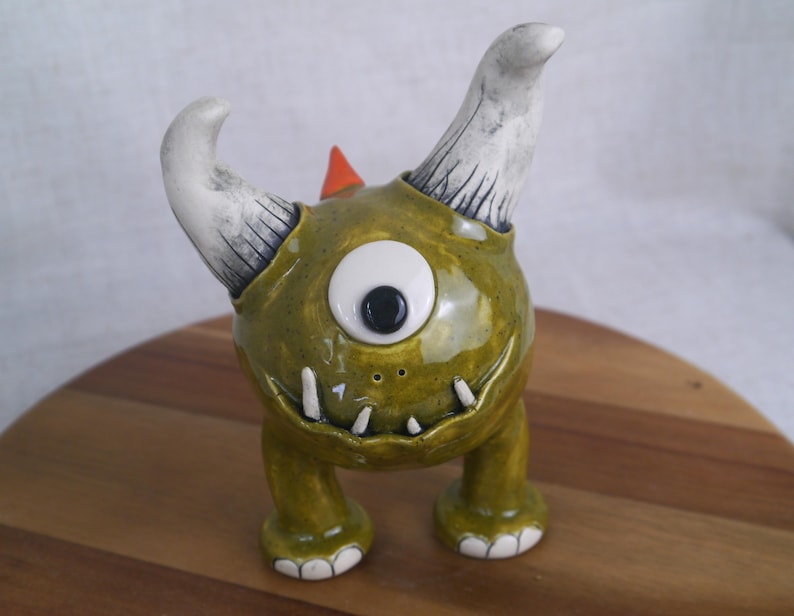 May include: A whimsical ceramic monster figurine in shades of olive green, white, and orange. The creature has one large eye, two curved horns, and a row of small teeth. The figurine is standing on a wooden surface.