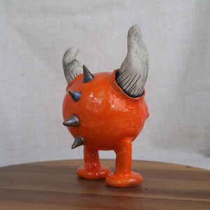 May include: A whimsical, orange ceramic sculpture of a creature with two gray horns and metallic spikes. The sculpture has a round body, two legs, and is set on a wooden surface.