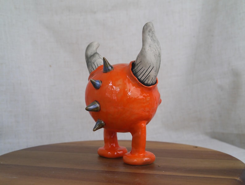 May include: A whimsical, orange ceramic sculpture of a creature with two gray horns and metallic spikes. The sculpture has a round body, two legs, and is set on a wooden surface.