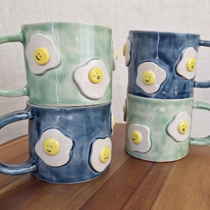 May include: Two sets of stacked ceramic mugs with a green and blue glaze. Each mug features a white, raised design of a fried egg with a smiley face.