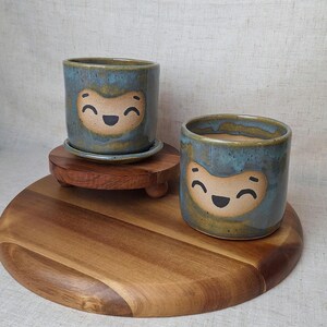 May include: Two ceramic planters with a smiling face design. The planters are cylindrical with a blue glaze and a tan face. One planter sits on a matching saucer, and both are on a wooden stand. The planters are suitable for small plants.