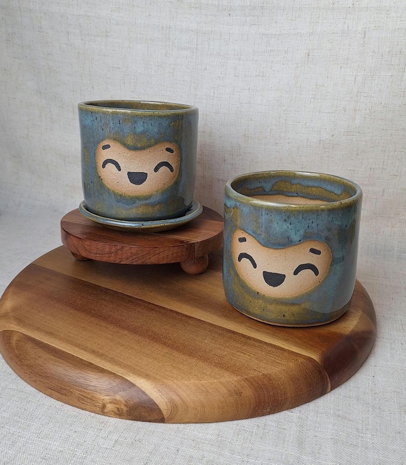 May include: Two ceramic planters with a smiling face design. The planters are cylindrical with a blue glaze and a tan face. One planter sits on a matching saucer, and both are on a wooden stand. The planters are suitable for small plants.