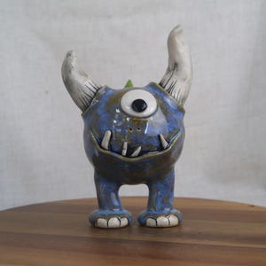 May include: A ceramic monster figurine with a blue body, one eye, and two white horns. The figurine has a toothy grin and small feet. The glaze gives it a glossy finish. The monster is standing on a wooden surface.