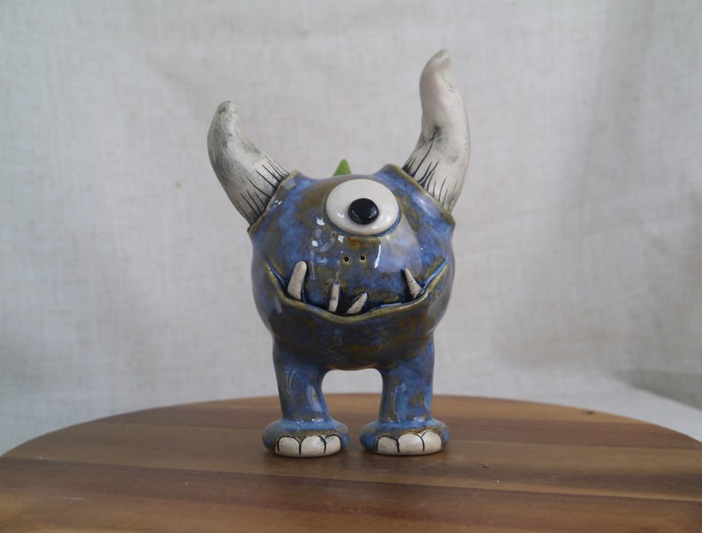 May include: A ceramic monster figurine with a blue body, one eye, and two white horns. The figurine has a toothy grin and small feet. The glaze gives it a glossy finish. The monster is standing on a wooden surface.
