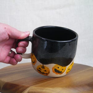 May include: Black and white ceramic mug with a black glaze and a white band around the bottom. The white band is decorated with orange and yellow jack-o'-lantern faces.