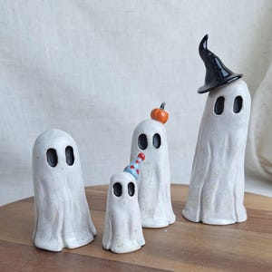 Könnte beinhalten: Vier weiße Keramik-Geisterfiguren mit schwarzen ovalen Augen. Ein Geist trägt einen schwarzen Hexenhut, ein anderer einen kleinen orangefarbenen Kürbis und ein dritter eine Partyhütchen. Die Geister stehen auf einer Holzoberfläche.