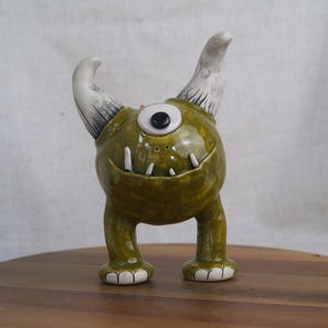 May include: A whimsical ceramic monster figurine with a single eye, two white horns with black detailing, and small white teeth. The figurine is primarily olive green with white accents on the feet. It stands on a wooden surface.