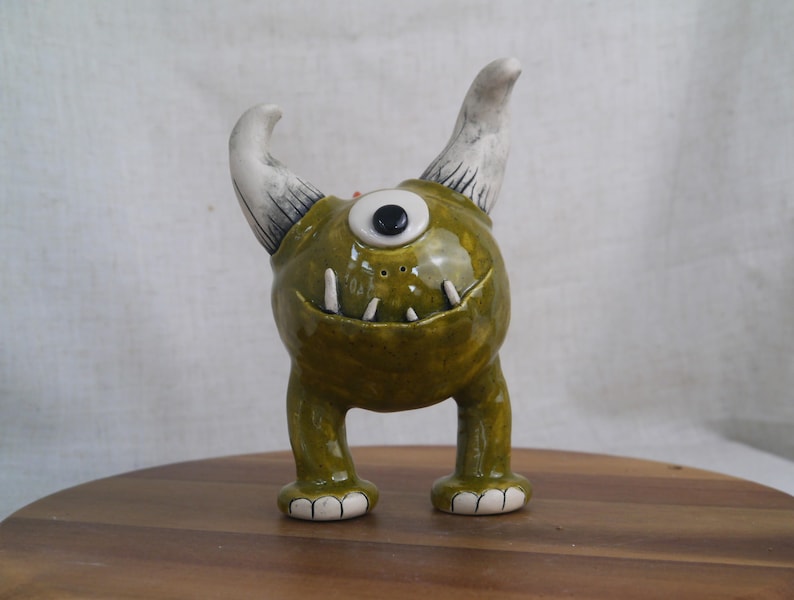 May include: A whimsical ceramic monster figurine with a single eye, two white horns with black detailing, and small white teeth. The figurine is primarily olive green with white accents on the feet. It stands on a wooden surface.