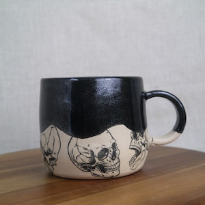 May include: Black ceramic mug with a white glaze and a black skull design. The mug has a rounded shape and a handle.