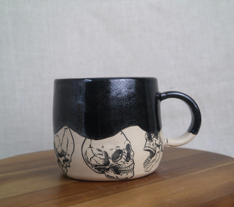 May include: Black ceramic mug with a white glaze and a black skull design. The mug has a rounded shape and a handle.