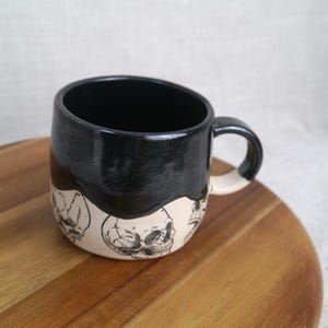 May include: A black ceramic mug with a white glaze and a skull design. The mug has a rounded shape and a handle.