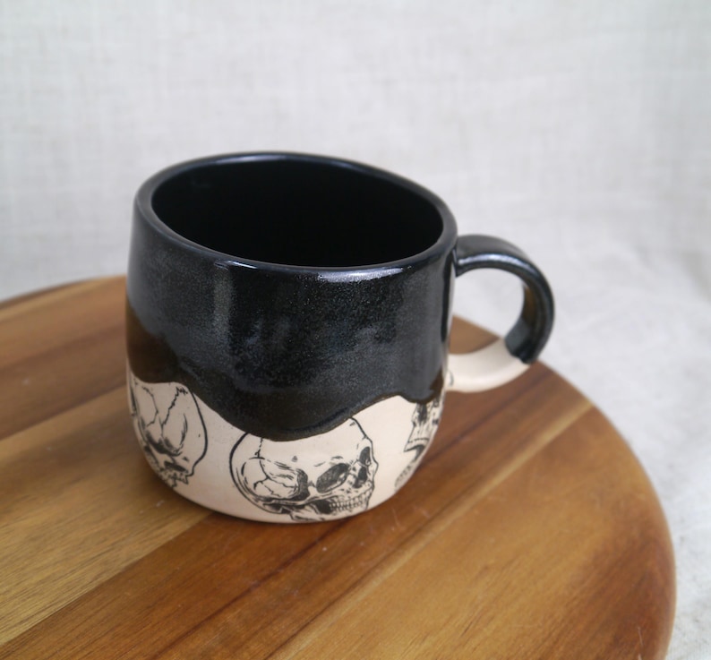May include: A black ceramic mug with a white glaze and a skull design. The mug has a rounded shape and a handle.