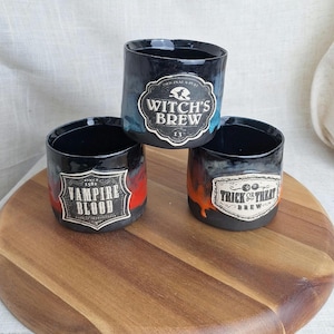 May include: Three black ceramic cups with labels reading "Witch's Brew", "Vampire Blood", and "Trick or Treat Brew". The cups have a glossy finish with colourful accents of blue, red, and orange. The cups are stacked on a wooden tray.