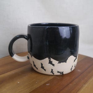 May include: A black ceramic mug with a white rim and handle. The mug has a black glaze and a white band with a black bat pattern.
