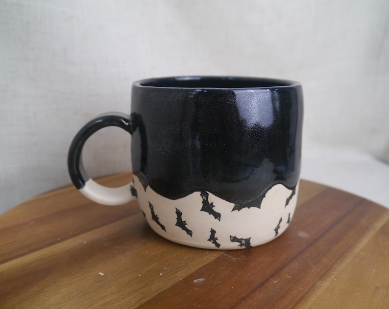 May include: A black ceramic mug with a white rim and handle. The mug has a black glaze and a white band with a black bat pattern.
