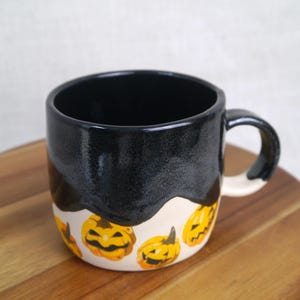 May include: A black ceramic mug with a white glaze and a black drip design. The mug is decorated with a pattern of yellow and orange jack-o'-lantern faces.
