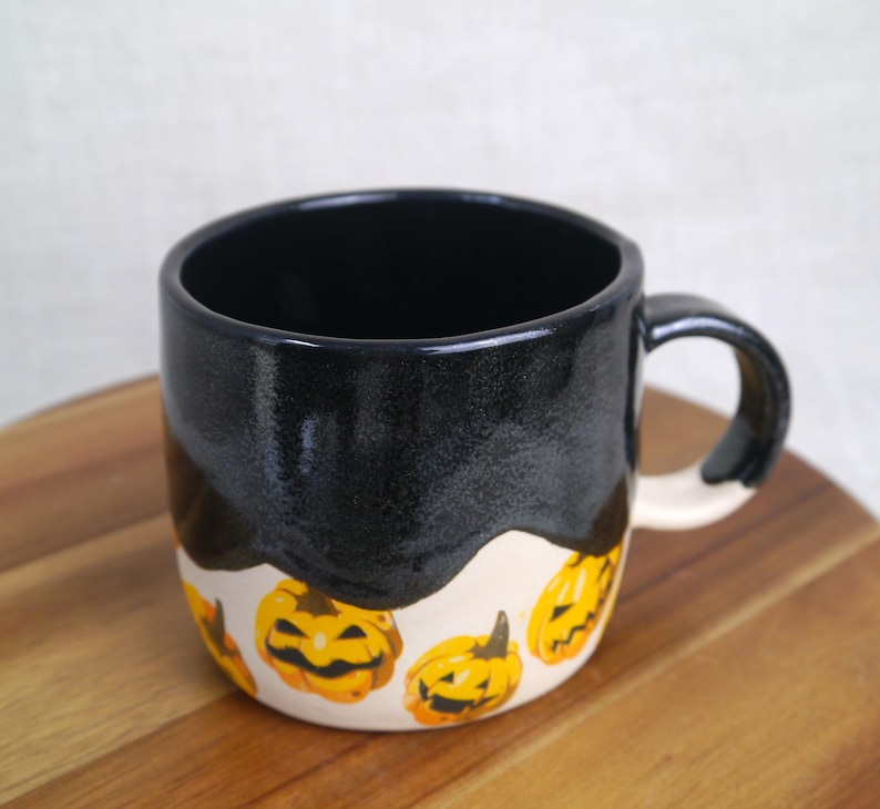 May include: A black ceramic mug with a white glaze and a black drip design. The mug is decorated with a pattern of yellow and orange jack-o'-lantern faces.