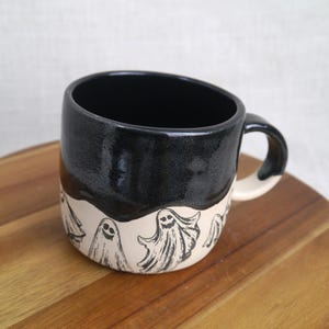 May include: A black and white ceramic mug with a black glaze on the top half and a white glaze on the bottom half. The bottom half features a design of four ghosts with smiling faces.