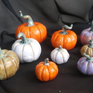 May include: A collection of ceramic pumpkins in various sizes and colors, including orange, white, purple, and tan. Each pumpkin has a green stem and is arranged on a dark background, perfect for fall or Halloween decor.