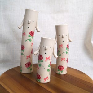 May include: Three quirky ceramic vases in cream, decorated with red roses and pink bows. Each vase features a face with black dot eyes and small, curved arms. They are displayed on a wooden surface.