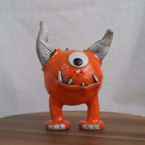 May include: A whimsical orange ceramic monster figurine with one large eye, two horns, and sharp teeth. The handmade creature has a glossy finish and stands on two short legs. A unique decorative item.