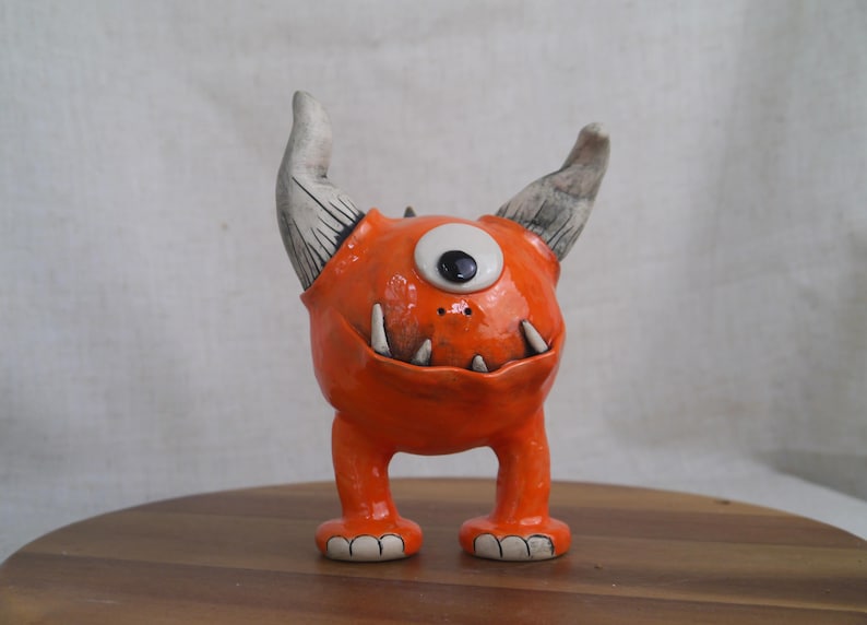 May include: A whimsical orange ceramic monster figurine with one large eye, two horns, and sharp teeth. The handmade creature has a glossy finish and stands on two short legs. A unique decorative item.