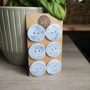 May include: Six pale blue ceramic buttons with a swirling design are presented on a brown card. Each button has two sewing holes. The card features a circular logo with the letters "M C". The buttons are on a light wooden surface.