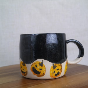 May include: Black ceramic mug with a white band and a Halloween theme. The band is decorated with orange and yellow jack-o'-lantern faces.