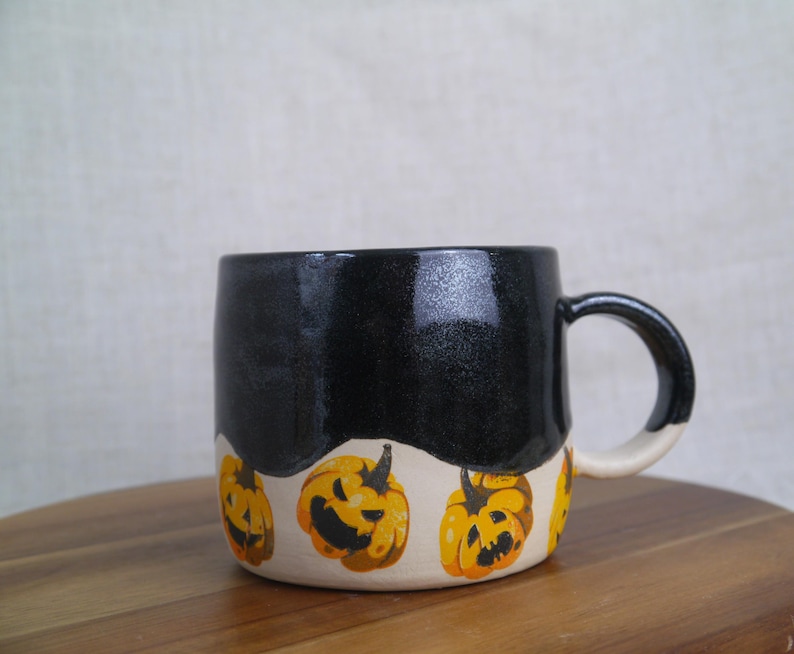 May include: Black ceramic mug with a white band and a Halloween theme. The band is decorated with orange and yellow jack-o'-lantern faces.